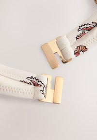 Fermoir métallique doré fixé à des sangles en tissu blanc avec des motifs floraux sur un fond clair.