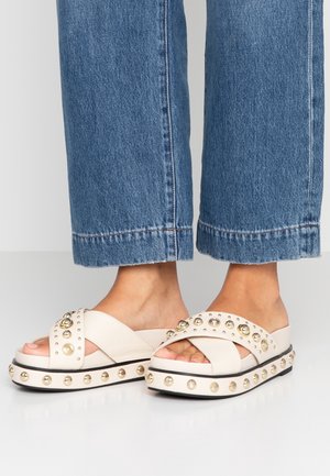 Stopy w białych sandałach na platformie typu slip-on z złotymi ćwiekami i szerokimi, krzyżującymi się paskami, zestawione z niebieskimi krótkimi jeansami.