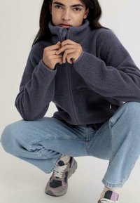 Giacca in fleece grigio scuro con zip e colletto alto, abbinata a jeans in denim azzurro chiaro e sneakers multicolori. Texture morbida e vestibilità comoda.