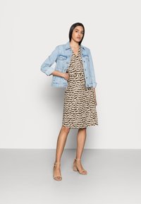 Jaqueta de ganga sobre um vestido castanho com padrões de linhas onduladas. Saltos bejes com tiras. Modelo em pé com uma mão no bolso contra um fundo simples.