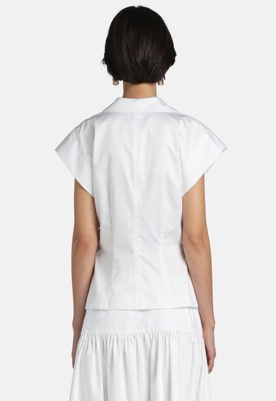 Femeie cu păr scurt și închis la culoare, purtând o bluză albă cu mâneci scurte și guler, și o fustă albă adunată, văzută din spate.