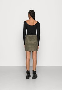 Top nero a maniche lunghe con scollatura off-the-shoulder, abbinato a una mini gonna in suede verde oliva con cerniera. Stivali neri alla caviglia completano il look.