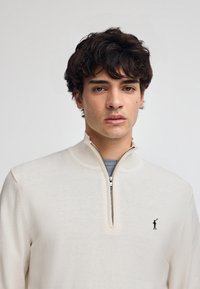 Krämfärgad zip-up tröja med hög krage, stickad struktur och en liten svart logotyp på bröstet. Modellen har kort krulligt hår.