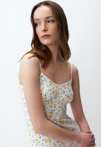 Blumenkleid mit dünnen Trägern, aus einem weißen Stoff, der mit kleinen blauen und gelben Blumen verziert ist. Eng anliegendes Oberteil mit einem leichten Volant.