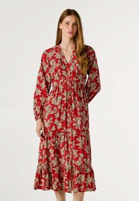 Robe florale rouge avec des manches longues, un décolleté en V et une taille froncée. Comprend une jupe à volants et un motif floral crème sur l'ensemble. Tissu lisse.