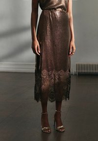 Frau trägt ein bronzefarbenes, glitzerndes Midikleid mit Spitzenaufsatz und goldenen High Heels, steht auf einem dunklen Holzboden vor einer grauen Wand.
