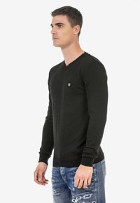 Schwarzer V-Ausschnitt-Pullover mit langen Ärmeln, feinem Strickmuster und kleinem Emblem auf der Brust. Kombiniert mit verwaschenen blauen Jeans.