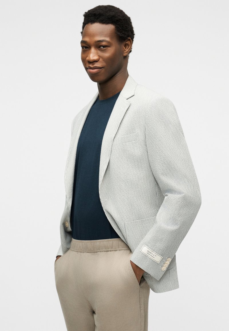 Giovane uomo che indossa un blazer leggero e testurizzato sopra una camicia blu navy con pantaloni beige, mani in tasca, guarda con sicurezza la fotocamera.