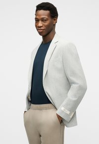 Jeune homme portant un blazer léger texturé sur une chemise bleu marine avec un pantalon beige, mains dans les poches, regardant la caméra avec confiance.