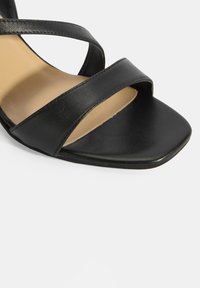 Zwarte leren sandaal met gekruiste banden, een vierkante neus en een gladde textuur met een beige binnenzool. Minimalistisch ontwerp, platte zool.