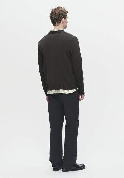 Suéter de punto verde oscuro con dobladillo y puños acanalados, superpuesto a una camisa de rayas verde claro y blanca, combinado con pantalones negros.