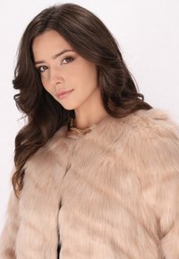 Faux fur coat in lichtbeige, met een zachte textuur en een volle kraag. Gecombineerd met een gouden ketting voor extra detail.