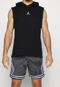 Hoodie senza maniche nera con logo bianco, abbinato a pantaloni corti in rete grigi con un motivo tie-dye e dettagli bianchi lungo i lati.