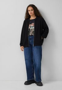 Veste noire zippée avec une finition texturée, associée à un t-shirt graphique et un jean bleu ample. Le look est complété par des chaussures noires.