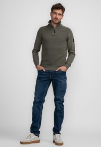 Olivgrüner, gerippter Zip-Hoodie mit hohem Kragen, kombiniert mit dunkelblauen Jeans und weißen Sneakers mit braunen Sohlen.