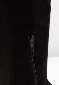 Černé semišové boty s vertikálním černým zipem na boku na plain bílém pozadí.