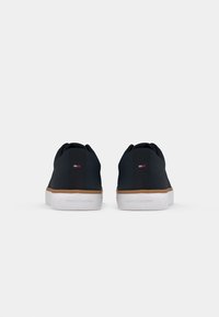 Et par sorte sneakers med hvide såler og brune detaljer, viser bagfra med lille Tommy Hilfiger logo på hælene og sålens branding.