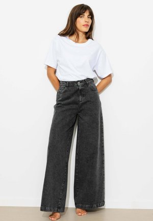 Femme aux cheveux bruns portant un t-shirt blanc ample rentré dans un jean noir taille haute à jambes larges, debout pieds nus contre un mur blanc.