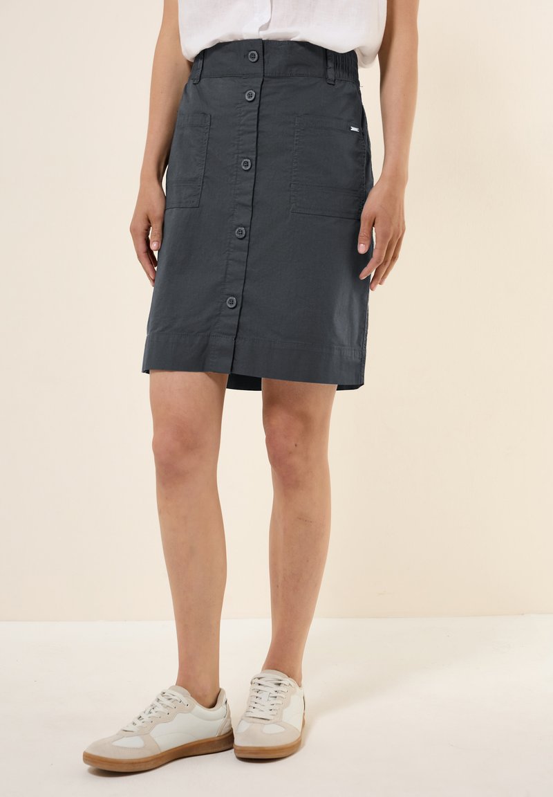Grauer knielanger Rock mit vorderen Knöpfen, zwei Seitentaschen und einem leicht strukturierten Stoff. Trägt weiße und beige Sneaker.