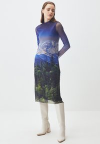 Abito lungo trasparente con maniche lunghe che presenta un design di paesaggio montano in blu e verde, abbinato a stivali bianchi al ginocchio con tacco a blocco.