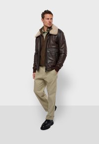 Chaqueta bomber de cuero marrón con cuello de forro polar, dos bolsillos frontales, combinada con un suéter oscuro con cierre y pantalones color crema. Botas negras.