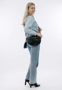 Zwarte leren crossbody tas met een ronde vorm, voorzien van een ritssluiting en een franje detail. Model draagt lichte spijkerbroek en zwarte hakken.