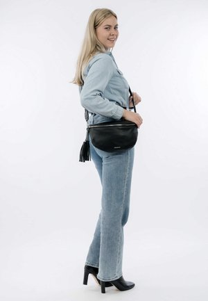 Black leather crossbody bag with a rounded shape, featuring a zip closure and tassel detail. Model wears light denim and black heels.