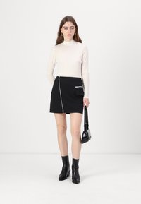 Gonna mini nera con zip, dotata di tasca laterale e dettaglio logo bianco, abbinata a una maglietta a maniche lunghe color crema e stivaletti neri alla caviglia.