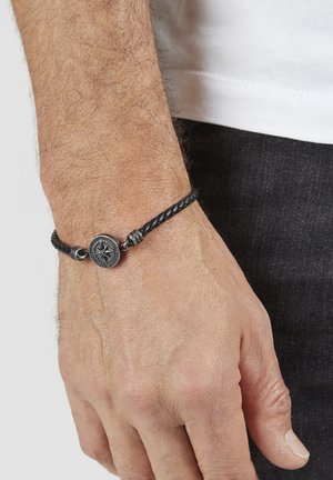 Schwarzes, geflochtenes Lederarmband mit einem runden Kompassanhänger in Silberoptik und metallischen Akzenten. Getragen an einem Handgelenk mit sichtbaren Haaren und Hautstruktur.