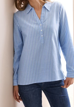 Vrouw die een lichtblauw, gestreept overhemd met lange mouwen en drie knopen draagt, gecombineerd met donkerblauwe spijkerbroek, staand tegen een muur.