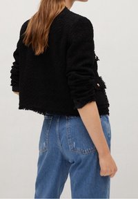 Musta lyhyt takki, jossa on teksturoitu kangas, repaleiset reunat ja nappikoristeet, yhdistettynä vaaleansinisiin korkeavyötäröisiin farkkuihin.