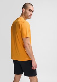 Orange kortärmad träningsskjorta i lätt material, med rund hals och diskret varumärke vid fållen. Bärs med svarta shorts.