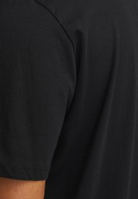 Juodas medvilninis T-shirt su apvalia iškirpte, trumpomis rankovėmis ir švelnia, lygia tekstūra. Siūlės detalė matoma ant peties.