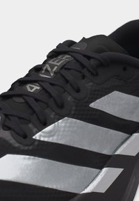 Κοντινό πλάνο μαύρου αθλητικού παπουτσιού Adidas με ασημένιες ρίγες και κείμενο "ADIZERO" στη γλώσσα, δείχνοντας τα κορδόνια και το υφασμάτινο επάνω μέρος με υφή.