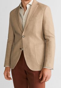 Muž oblečený v béžovém saku přes světle béžovou košili a hnědé kalhoty, stojící s uvolněnýma rukama podél těla.