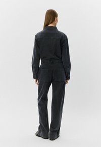 Sötét farmer overál laza szabással, hosszú ujjakkal és felhajtott szegélyekkel. Nagy hátsó zsekei és összehúzott dereka van. Minimalista dizájn.