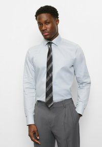 Hellblaues Hemd mit Kragen, kombiniert mit grauen, plissierten Hosen. Eine schwarze Krawatte mit weißen diagonalen Streifen sorgt für Kontrast.