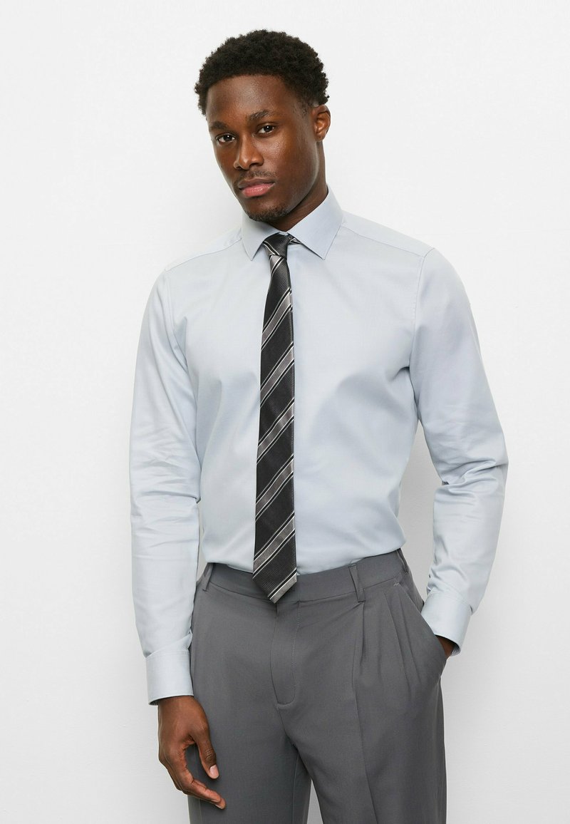 Hellblaues Hemd mit Kragen, kombiniert mit grauen, plissierten Hosen. Eine schwarze Krawatte mit weißen diagonalen Streifen sorgt für Kontrast.