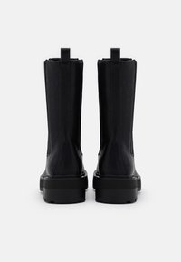 Botines de cuero negro con textura lisa, forma rectangular y suelas gruesas. Incluyen tiradores traseros y detalles de costura mínimos.