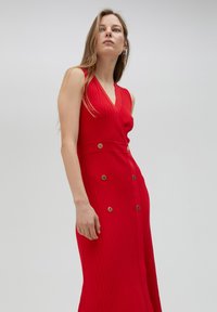 Rode geribbelde maxi-jurk met een V-hals, een aansluitende taille en gouden knoopaccenten aan de voorkant. De stof heeft een gestructureerde afwerking.