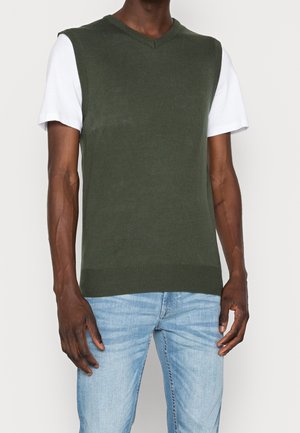 Mand iført en mørkegrøn ærmeløs V-hals sweater over en hvid kortærmet T-shirt og lyseblå jeans.
