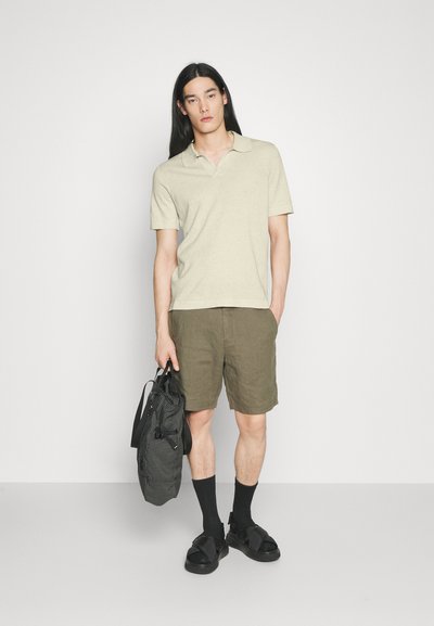 Uomo con capelli lunghi neri che indossa una polo beige, pantaloncini oliva, calzini neri con sandali, tiene una borsa nera con motivo, in piedi contro uno sfondo bianco.
