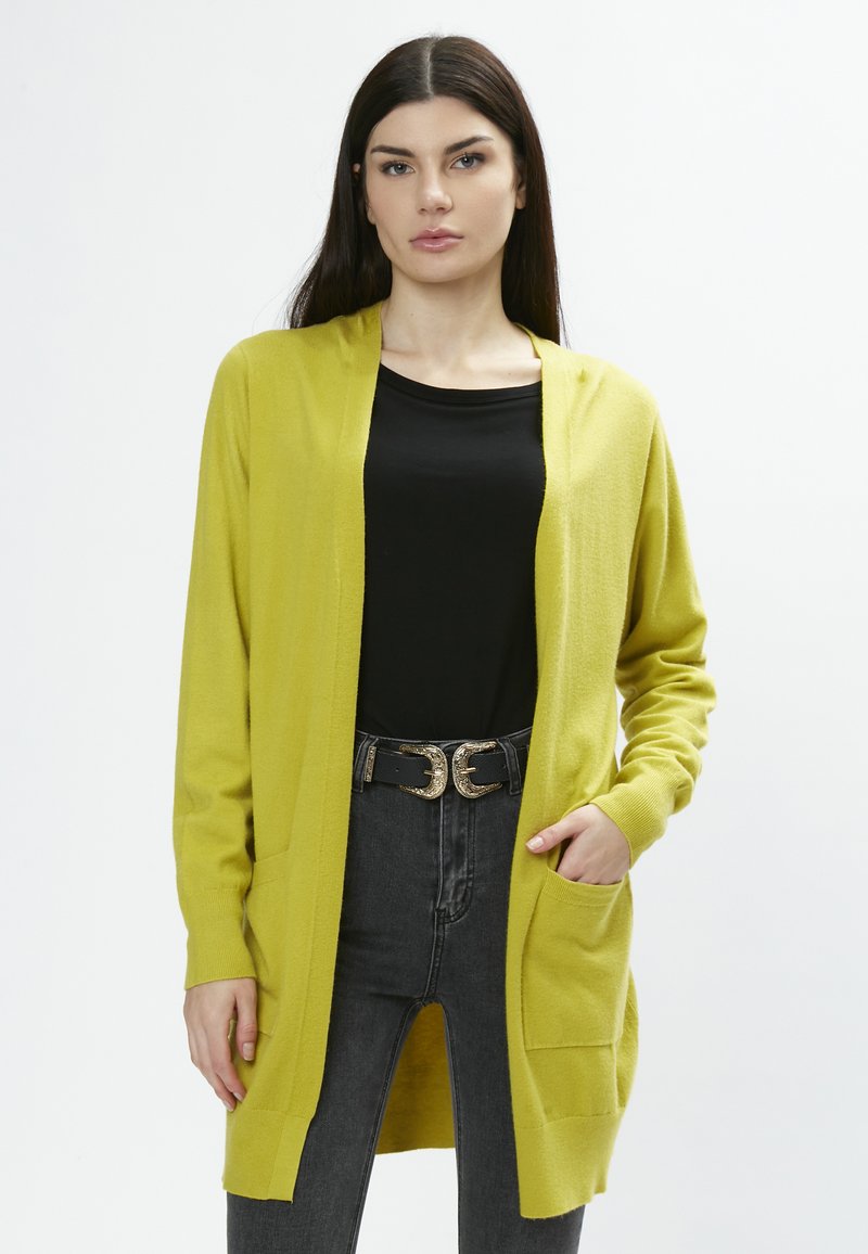 Cardigan jaune en tricot doux, avec des manches longues, une fermeture ouverte et deux poches latérales. Porté sur un haut noir et un jean taille haute.