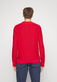 Suéter rojo de punto con cuello redondo, mangas largas y dobladillo acanalado. Tejido texturizado con un patrón sutil. Combinado con jeans azules.