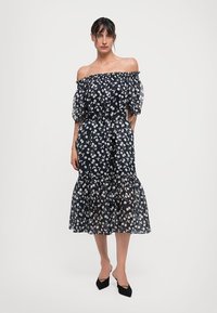 Marineblaues Off-Shoulder-Kleid mit weißem Blumenmuster, durchsichtigen Puffärmeln, taillierter Taille und gestuftem Rüschensaum. Schwarze spitze Schuhe.