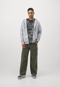 Sivá mikina na zips s kapucňou vrstvená nad čiernou a béžovou tričkou s tie-dye vzorom. Olivové cargo nohavice so bočnými vreckami. Sivé športové topánky s priehľadnými podrážkami.