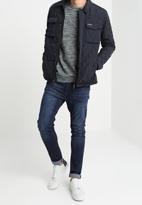 Chaqueta de nylon acolchada en azul marino con dos bolsillos en el pecho, suéter gris, jeans oscuros y zapatillas blancas. La chaqueta presenta un cierre con cremallera y un branding sutil.