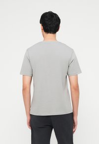 Άνδρας με κοντά μαύρα μαλλιά, που φοράει απλό γκρι ανοιχτό κοντομάνικο μπλουζάκι και μαύρο παντελόνι, στέκεται γυρισμένος με την πλάτη, μπροστά σε λευκό φόντο.