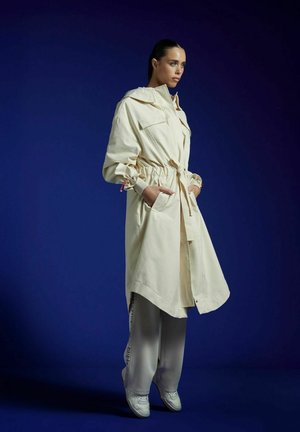 Donna con cappotto lungo beige con cappuccio e coulisse in vita, pantaloni ampi grigio chiaro e scarpe da ginnastica bianche, in piedi davanti a uno sfondo blu scuro.