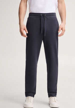 Man draagt donkerblauwe joggingbroek met trekkoord en witte sneakers, staand tegen een lichte achtergrond.